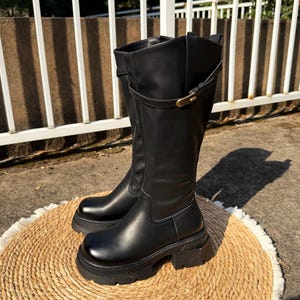May include: A pair of black knee-high boots with a chunky sole. The boots have a smooth leather-like finish, a strap detail with a gold-colored buckle, and a rounded toe. They are placed on a round, woven mat.