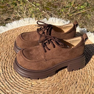 Könnte beinhalten: Ein Paar braune Oxford-Schuhe aus Wildleder mit dicken Profilsohlen. Die Schuhe haben eine runde Spitze, einen Schnürverschluss und eine Zuglasche an der Ferse. Sie werden auf einer gewebten Matte präsentiert.