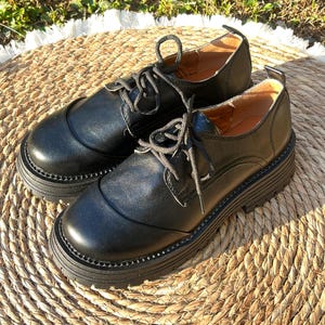 May include: A pair of black leather Oxford shoes with thick, lug soles. The shoes have a classic lace-up design and a rounded toe. The soles are approximately 2 inches thick. The shoes are displayed on a woven, circular mat.
