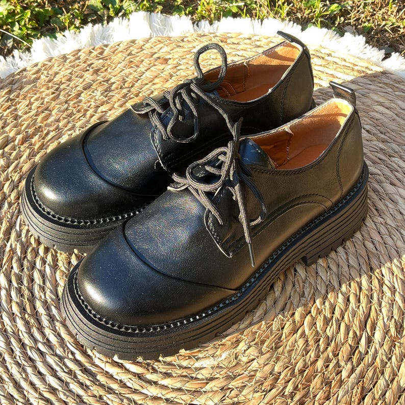 May include: A pair of black leather Oxford shoes with thick, lug soles. The shoes have a classic lace-up design and a rounded toe. The soles are approximately 2 inches thick. The shoes are displayed on a woven, circular mat.