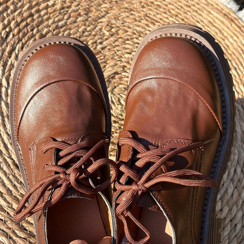 May include: A pair of brown leather oxford shoes with matching laces. The shoes have a rounded toe and a thick, textured sole. The shoes are displayed on a woven, circular surface.
