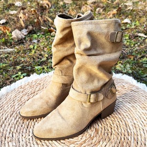 Puede incluir: Un par de botas de ante color canela, de media caña. Las botas presentan un diseño holgado, hebillas decorativas y un tacón bajo. Las botas se exhiben sobre una estera tejida.