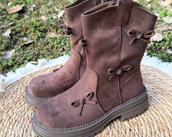 May include: A pair of brown suede boots with a platform sole. The boots have a mid-calf height and are adorned with small bow accents. The boots are displayed on a woven mat, showcasing their design and texture.