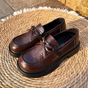 May include: A pair of brown leather loafers with bow detailing. The shoes have a rounded toe and a thick black sole. The loafers are on a woven mat.