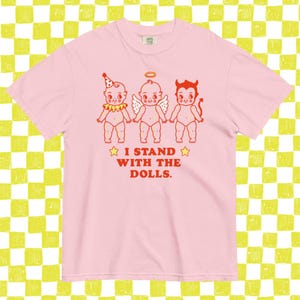 May include: Pink t-shirt featuring a graphic of three cartoon babies: one with a party hat, one with a halo and wings, and one with devil horns and a tail. The text "I STAND WITH THE DOLLS" is printed below the figures in red.