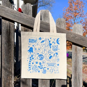 May include: A white tote bag with blue illustrations of a monster, a unicorn, a tiger, a snake, a baby, a palm tree, a sheep, a banana, a cherry, a mushroom, a moon, a star, and a diamond. The text "Bubble Wrapp" and "Tigersheep Friends" is printed on the bag.