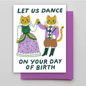 May include: A greeting card with the text "LET US DANCE ON YOUR DAY OF BIRTH" features two anthropomorphic cats in festive attire. One cat wears a green and purple dress, the other a purple jacket and brown shorts. The card is paired with a purple envelope.