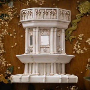 May include: A white, architectural wall-mounted fountain sculpture featuring a multi-tiered design with vertical columns, arched window-like cutouts, and a wide basin. The structure has a weathered, off-white finish with subtle brown detailing.