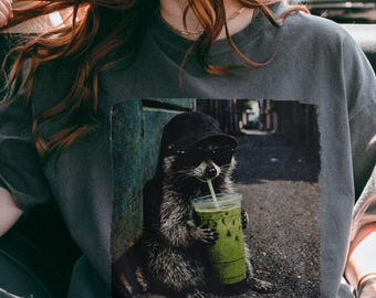 Camiseta con diseño de mapache bebiendo matcha latte helado, con colores reconfortantes y meme animal.
