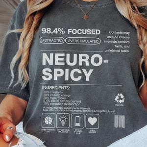 Op de afbeelding: Donkergrijs t-shirt met de witte tekst "NEURO-SPICY". Het shirt bevat tekst over focus, afleiding en overprikkeling, evenals een lijst met "INGREDIËNTEN". Het heeft ook iconen en een recyclingsymbool.