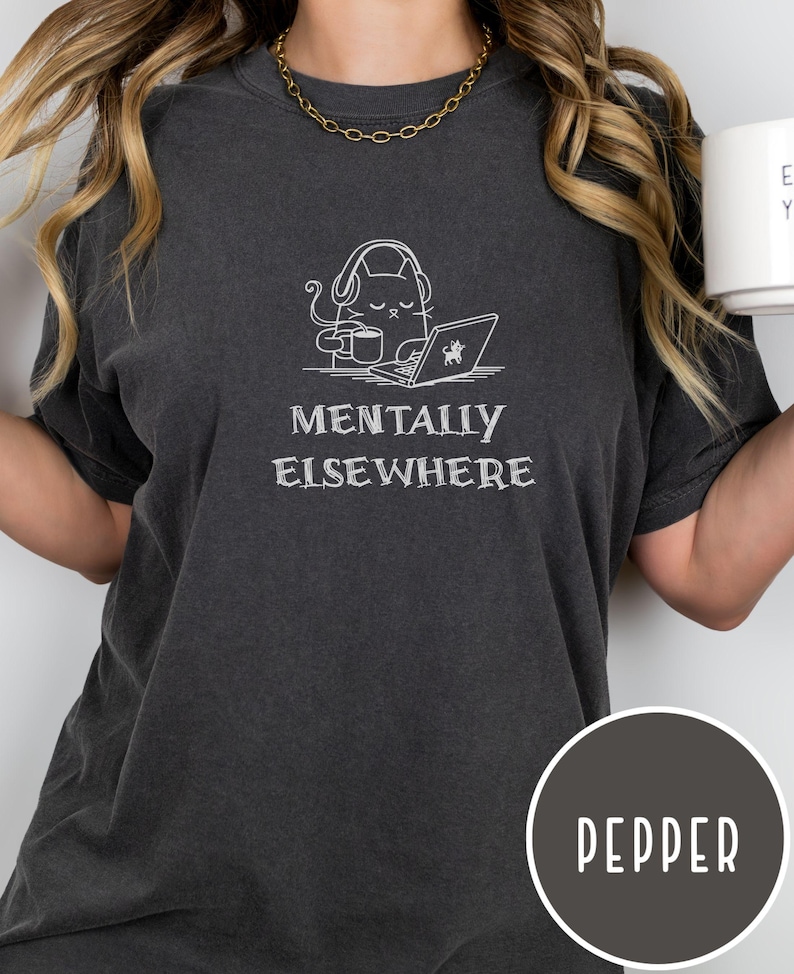 May include: Dark gray t-shirt with the phrase "MENTALLY ELSEWHERE" and a graphic of a cat wearing headphones, using a laptop, and holding a mug. The shirt is a pepper color.