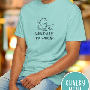 May include: A light blue t-shirt with the text "MENTALLY ELSEWHERE" and a graphic of a cat wearing headphones, using a laptop, and drinking coffee. The shirt is a solid color and has short sleeves.