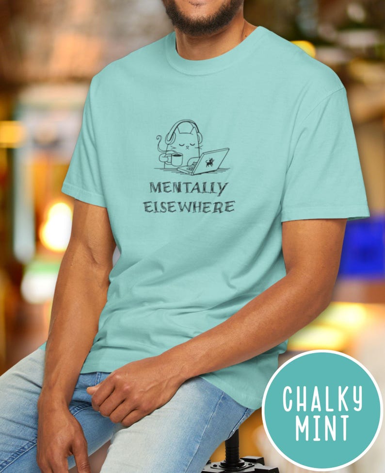 May include: A light blue t-shirt with the text "MENTALLY ELSEWHERE" and a graphic of a cat wearing headphones, using a laptop, and drinking coffee. The shirt is a solid color and has short sleeves.