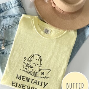 May include: A light yellow t-shirt with the words "MENTALLY ELSEWHERE" and a graphic of a cat wearing headphones, holding a coffee cup, and using a laptop. A tan hat and blue jeans are also in the image.