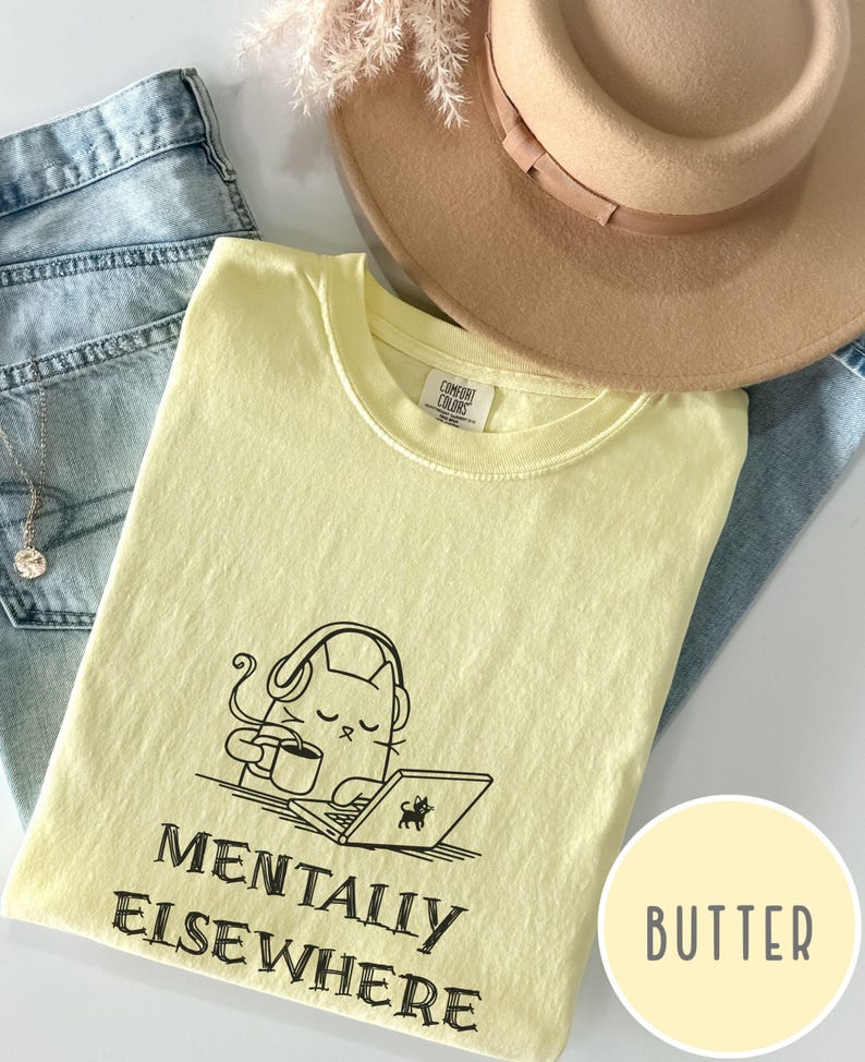 May include: A light yellow t-shirt with the words "MENTALLY ELSEWHERE" and a graphic of a cat wearing headphones, holding a coffee cup, and using a laptop. A tan hat and blue jeans are also in the image.