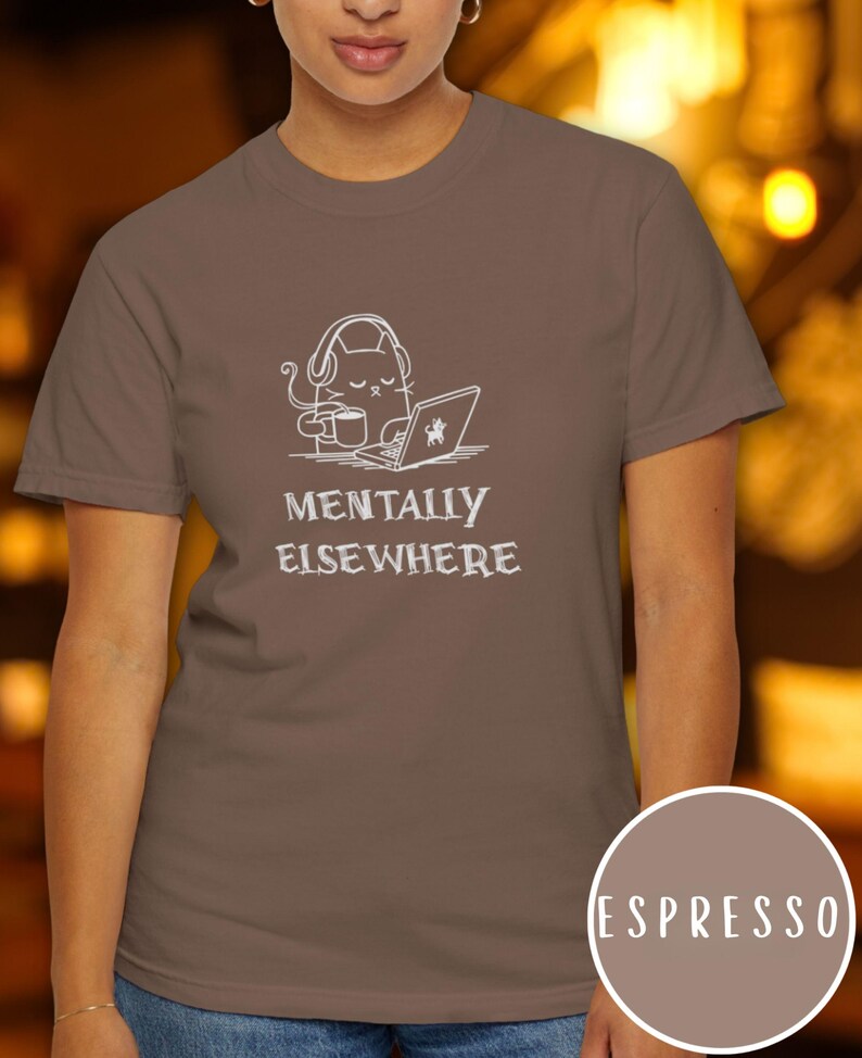 May include: Espresso-colored t-shirt featuring a white graphic of a cat wearing headphones, holding a coffee cup, and using a laptop. The text "MENTALLY ELSEWHERE" is below the graphic. A circular color swatch reads "ESPRESSO".