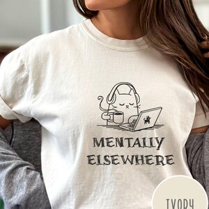 May include: Ivory-colored t-shirt featuring a black and white illustration of a cat wearing headphones, holding a coffee cup, and using a laptop. The text "MENTALLY ELSEWHERE" is below the graphic. The shirt is a light cream color.