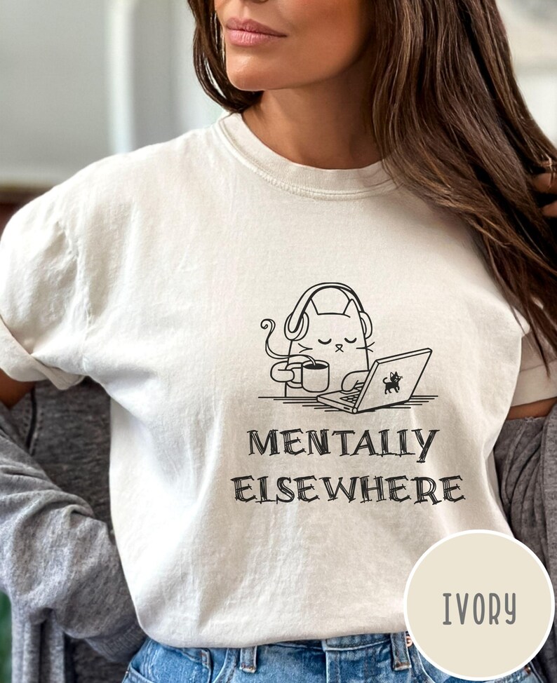 May include: Ivory-colored t-shirt featuring a black and white illustration of a cat wearing headphones, holding a coffee cup, and using a laptop. The text "MENTALLY ELSEWHERE" is below the graphic. The shirt is a light cream color.