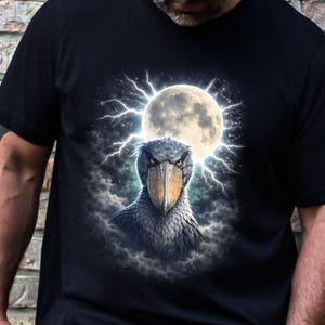Op de afbeelding: Zwart t-shirt met een gedetailleerde illustratie van een schoenbekooievaar, een volle maan en bliksem. De vogel heeft een grote snavel en een intense blik. Het ontwerp is gecentreerd op het t-shirt.