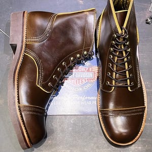 May include: A pair of brown leather boots with yellow stitching and laces. The boots have a cap toe and a thick sole. The boots are made by Harley-Davidson.