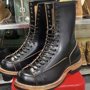Puede incluir: Par de botas de trabajo de cuero negro con diseño de cordones. Las botas tienen una silueta alta, una puntera redondeada y una suela marrón claro. Las botas se exhiben sobre una superficie roja, con otras botas visibles en el fondo.