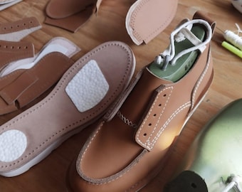 May include: A close-up of a brown leather shoe with white stitching. The shoe is on a wooden surface with various leather pieces and tools for shoemaking.
