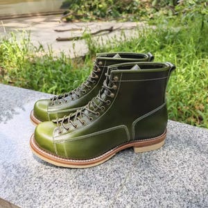 Puede incluir: Un par de botas de cuero verde oliva con diseño de cordones. Las botas tienen punta redondeada, suela marrón claro y costuras blancas. Las botas están sobre una superficie de piedra gris.