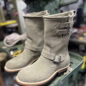 May include: A pair of light green suede boots with multiple buckle fastenings. The boots have a rounded toe and a light-coloured sole. The boots are on a green surface.
