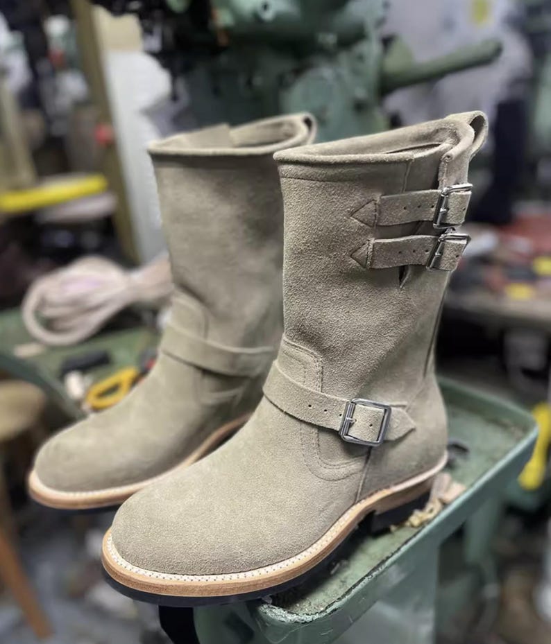 May include: A pair of light green suede boots with multiple buckle fastenings. The boots have a rounded toe and a light-coloured sole. The boots are on a green surface.
