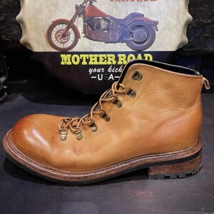 May include: A tan leather boot with a rounded toe and metal eyelets for the laces. The boot has a dark brown sole and heel. The background features a vintage sign with a motorcycle and the words "MOTHER ROAD - USA".