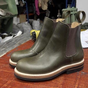 May include: Olive green leather Chelsea boots with rounded toes, elastic side panels, and pull tabs. The soles feature a layered design with light brown and black sections. The boots are presented on a wooden surface. Approx. 25 cm in length.