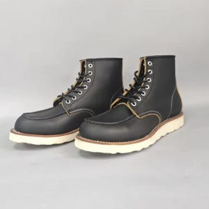 Puede incluir: Un par de botas de trabajo de cuero negro con suelas blancas y costuras color canela. Las botas tienen un diseño con cordones con ojales metálicos y una puntera redondeada. Las botas son nuevas y sin usar.
