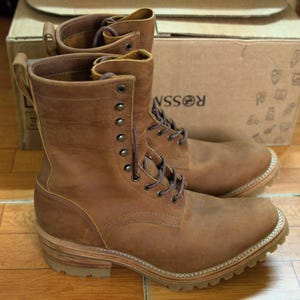 Könnte beinhalten: Ein Paar wadenhohe braune Lederstiefel mit Schnürung und dunklen Metallösen, einer Zugschlaufe an der Ferse, einem geschichteten Holzabsatz und einer dicken, profilierten Gummisohle mit sichtbaren weißen Nähten entlang des Rahmenbesatzes.