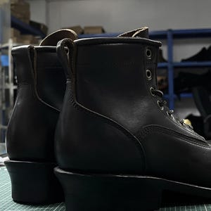 Op de afbeelding: Close-up van twee zwarte leren werklaarzen. De laarzen hebben een vetersluiting met metalen oogjes, een ronde neus en een dikke hak. Het leer lijkt glad en gepolijst, met zichtbare stikdetails. De achtergrond is wazig.