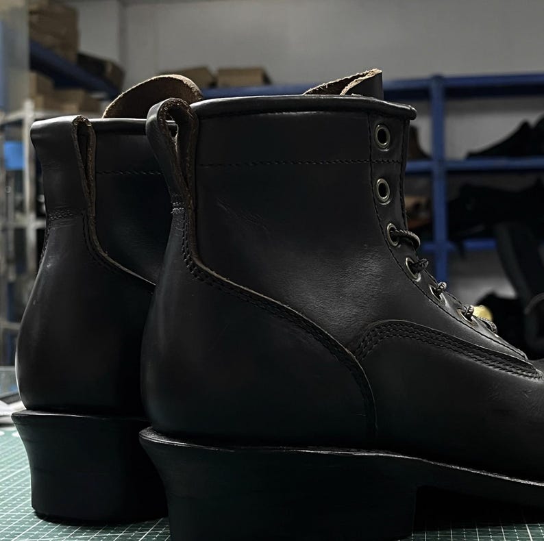 Op de afbeelding: Close-up van twee zwarte leren werklaarzen. De laarzen hebben een vetersluiting met metalen oogjes, een ronde neus en een dikke hak. Het leer lijkt glad en gepolijst, met zichtbare stikdetails. De achtergrond is wazig.