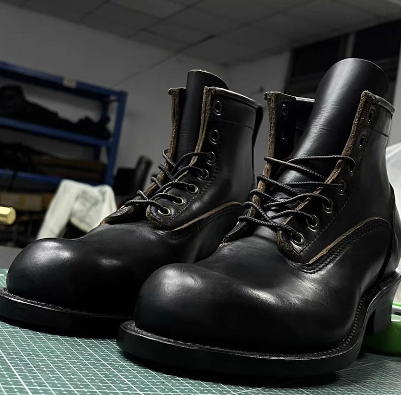 Op de afbeelding: Een paar zwarte leren werklaarzen met een vetersluiting. De laarzen hebben een ronde neus, een dikke zool en een stevige hak. Het leer lijkt versleten, waardoor de laarzen een stoere uitstraling hebben. De laarzen staan op een groene ondergrond.