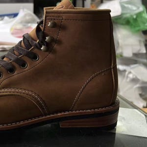 May include: A close-up of a brown leather boot with dark brown laces and metal eyelets. The boot features a sturdy sole and visible stitching details. The leather has a textured appearance, suggesting durability and quality.