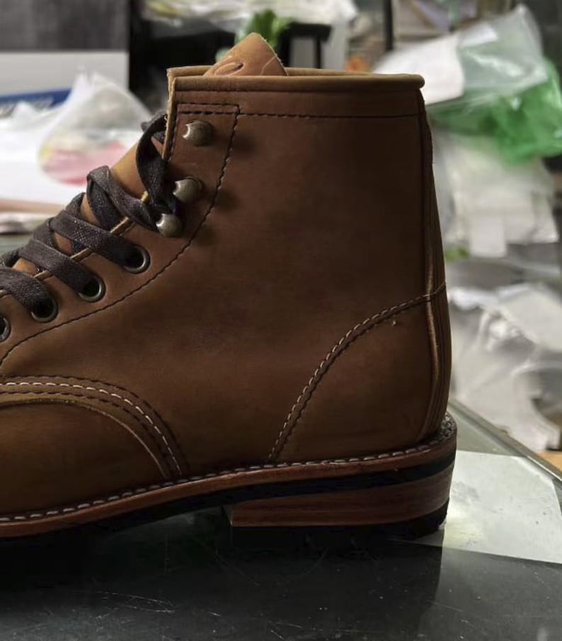 May include: A close-up of a brown leather boot with dark brown laces and metal eyelets. The boot features a sturdy sole and visible stitching details. The leather has a textured appearance, suggesting durability and quality.