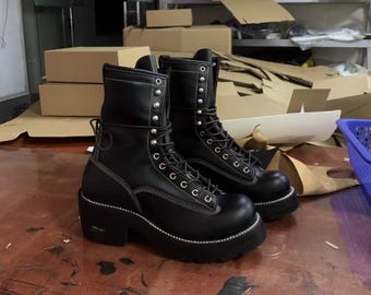 Könnte beinhalten: Ein Paar schwarze Leder-Kampfstiefel mit weißen Nähten und silberfarbenen Ösen. Die Stiefel haben eine klobige Plateausohle und sind vorne geschnürt. Die Stiefel stehen auf einer Holzoberfläche.