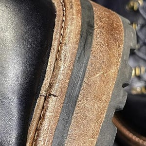 May include: Close-up of a black leather boot with a brown and black sole. The sole has a textured, rugged design. The stitching is visible along the side of the boot, and the leather appears worn.