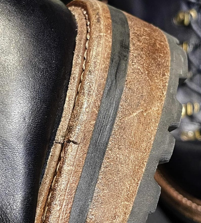 May include: Close-up of a black leather boot with a brown and black sole. The sole has a textured, rugged design. The stitching is visible along the side of the boot, and the leather appears worn.