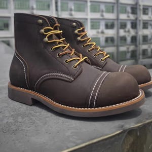 May include: A pair of brown leather work boots with yellow laces. The boots have a cap toe design and white stitching accents. The soles are brown with a light brown trim. The boots are on a grey surface.