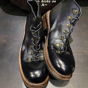 May include: A pair of black leather boots with a lace-up design and brass-colored eyelets. The boots have a brown sole and a sturdy, rugged appearance, suitable for outdoor activities. The boots are displayed on a dark surface.