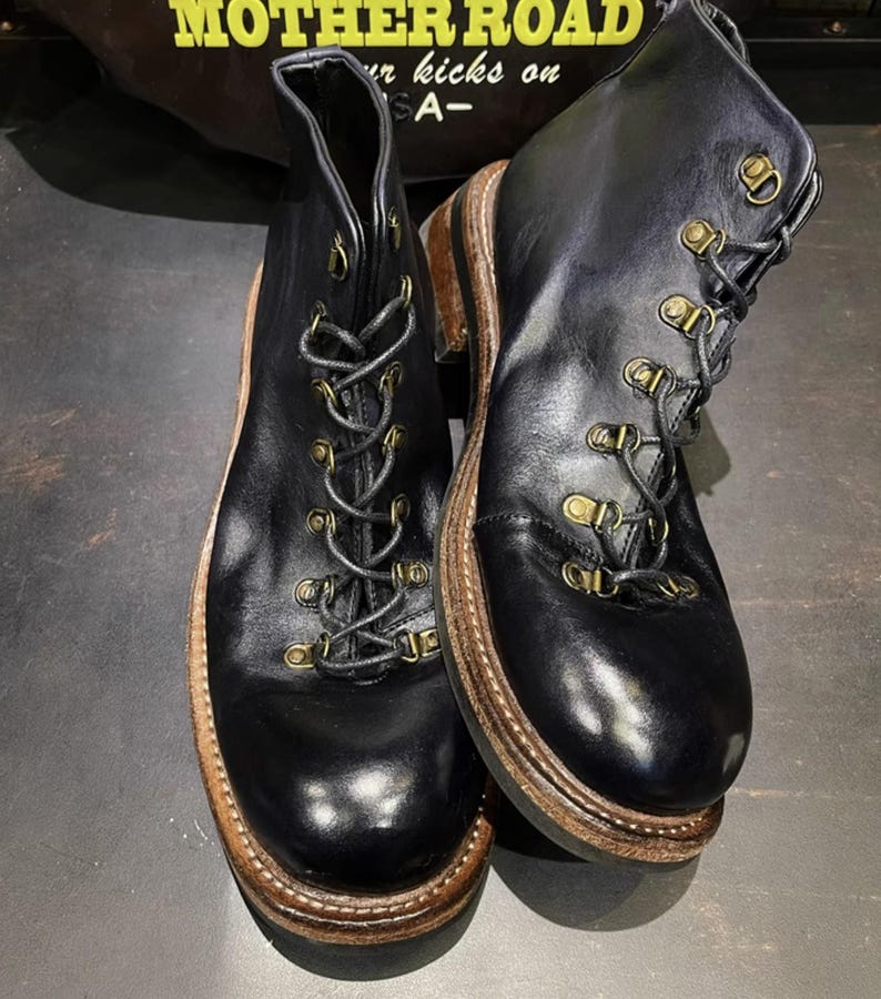 May include: A pair of black leather boots with a lace-up design and brass-colored eyelets. The boots have a brown sole and a sturdy, rugged appearance, suitable for outdoor activities. The boots are displayed on a dark surface.