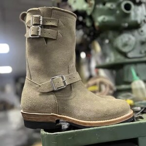 May include: A tan suede boot with three silver buckles. The boot has a rounded toe and a stacked heel. The boot is a mid-calf height and has a rugged, vintage look. The boot is likely made for motorcycle riding or work.