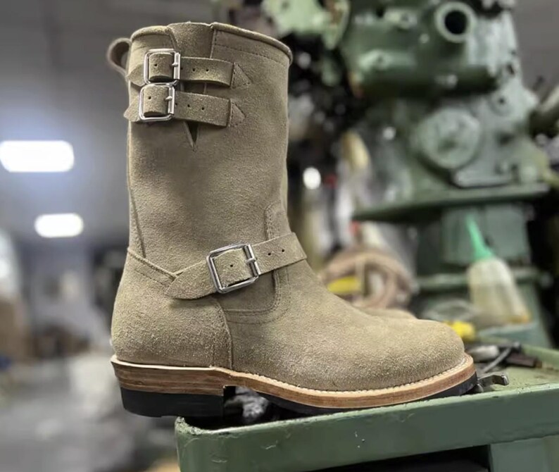 May include: A tan suede boot with three silver buckles. The boot has a rounded toe and a stacked heel. The boot is a mid-calf height and has a rugged, vintage look. The boot is likely made for motorcycle riding or work.