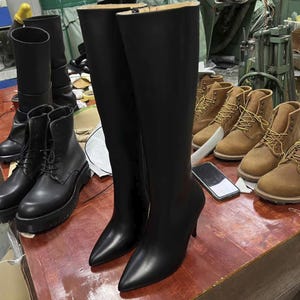 Puede incluir: Un par de botas negras de cuero hasta la rodilla con punta y tacones de aguja. Tambi&eacute;n se ven varios pares de botas de cuero negras y marrones, lo que sugiere un taller o exhibici&oacute;n de calzado.