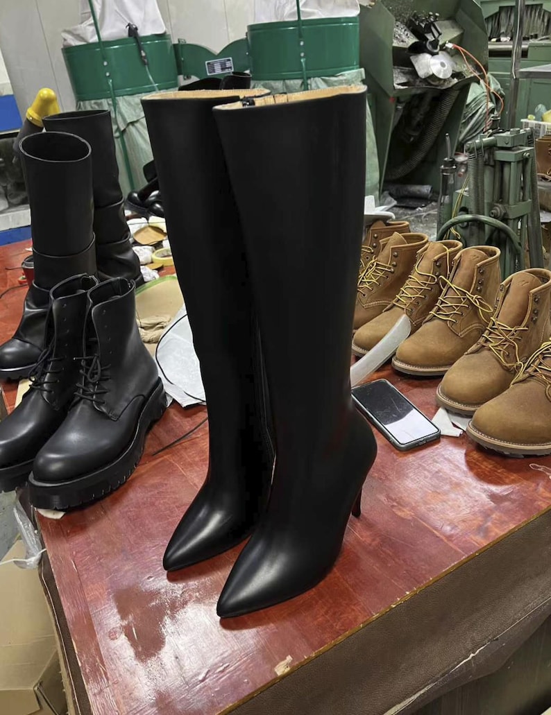 Puede incluir: Un par de botas negras de cuero hasta la rodilla con punta y tacones de aguja. Tambi&eacute;n se ven varios pares de botas de cuero negras y marrones, lo que sugiere un taller o exhibici&oacute;n de calzado.
