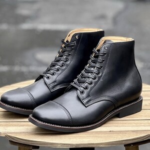 May include: A pair of black leather boots with cap toes and lace-up fastenings. The boots are presented on a wooden table, highlighting their polished finish and classic design. The boots have a sturdy sole and a high-top silhouette.