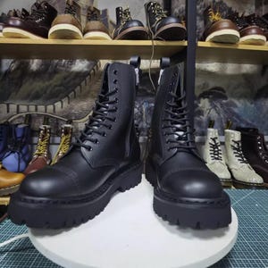 May include: Black leather combat boots with a lace-up design and side zippers. The boots have a thick, lugged sole and are displayed on a white surface. Other boots are visible on shelves in the background.